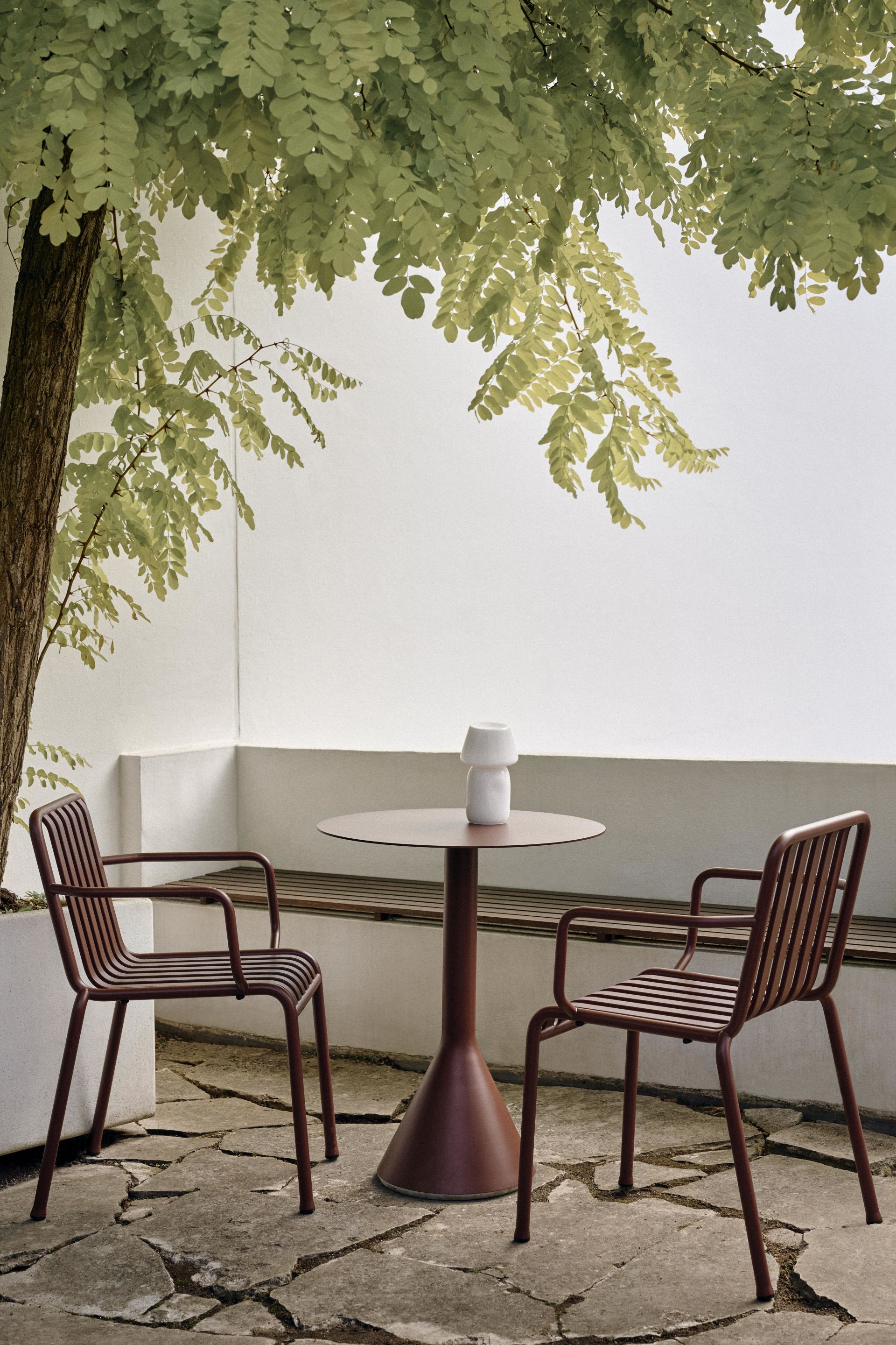 Der HAY Palissade Cone Tisch in rot mit zwei Palissade Dining Stühlen auf einer Terrasse unter einem Baum. 
